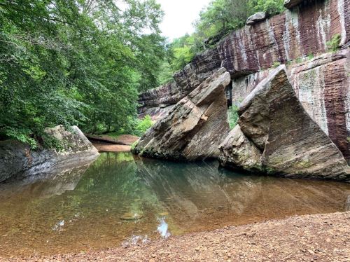A serene natural scene featuring large rock formations beside a calm, reflective pool of water surrounded by greenery.
