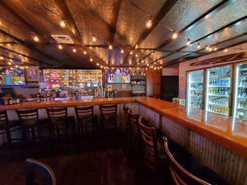 A cozy bar interior with a wooden counter, string lights, and a well-stocked fridge, featuring TVs showing sports.