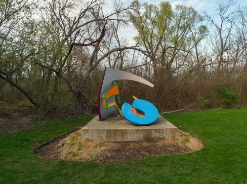 Colorful abstract sculpture on a stone base, surrounded by trees and grass in a natural setting.