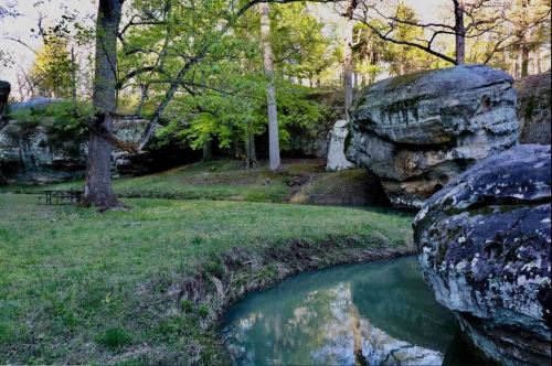 A serene landscape featuring a creek, lush greenery, and large rock formations under a clear sky.