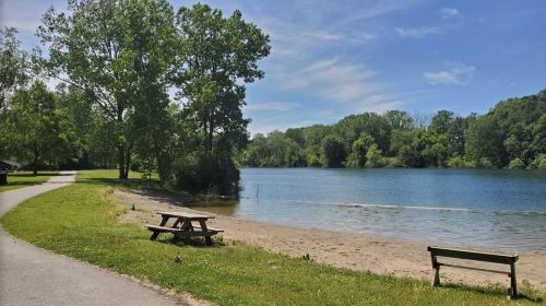 A serene lakeside scene with green trees, a sandy beach, and picnic tables along a walking path under a clear blue sky.