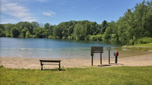 A serene lake with a sandy beach, surrounded by trees, featuring signs and a bench in the foreground under a clear blue sky.