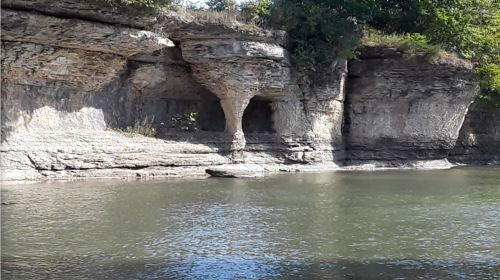Rocky cliff formation beside a calm river, with greenery above and a small cave-like opening in the stone.