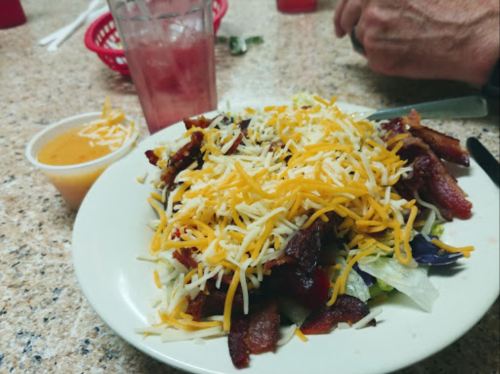 A plate of salad topped with bacon and shredded cheese, alongside a cup of dressing and a glass of pink drink.