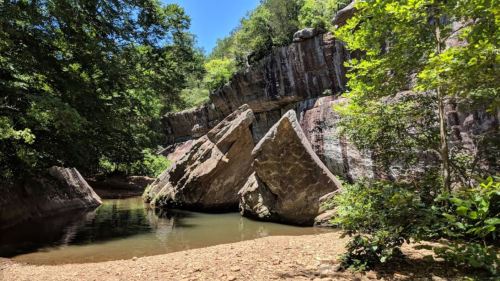 A serene landscape featuring large rocks by a calm water body, surrounded by lush greenery and steep cliffs.