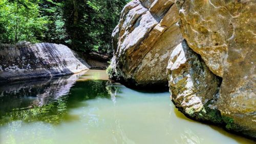 A serene river flows between large rocky cliffs, surrounded by lush greenery and reflecting the sunlight.