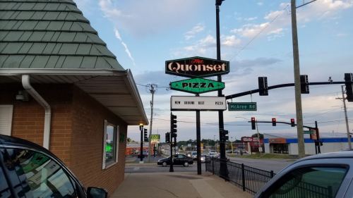 Sign for "The Quonset Pizza" with a new menu item announcement, viewed from the street near traffic lights.