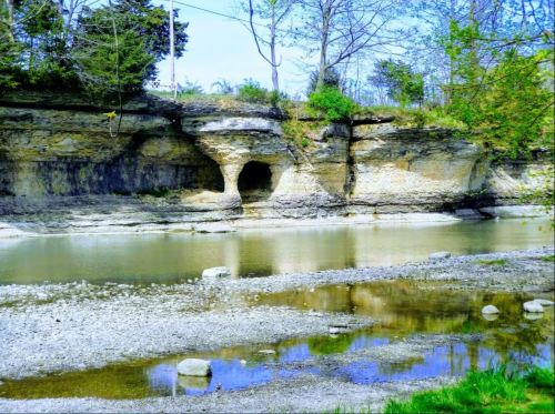 A serene river scene with rocky cliffs, trees, and a cave entrance, reflecting in the calm water under a clear blue sky.
