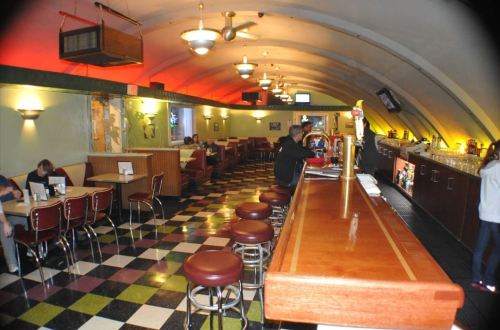 A retro diner interior with checkered floors, booths, and a long bar, featuring warm lighting and a cozy atmosphere.