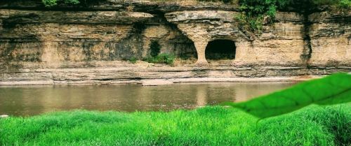 A serene river scene with a rocky cliff featuring a cave, surrounded by lush green grass and trees.