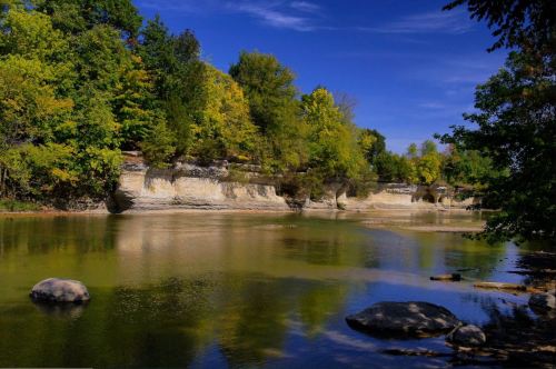 A serene river scene with rocky banks, lush green trees, and a clear blue sky reflecting on the water's surface.