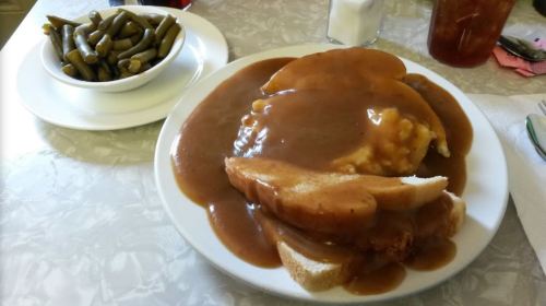 A plate of sliced meat covered in brown gravy, served with mashed potatoes and toast, alongside a bowl of green beans.