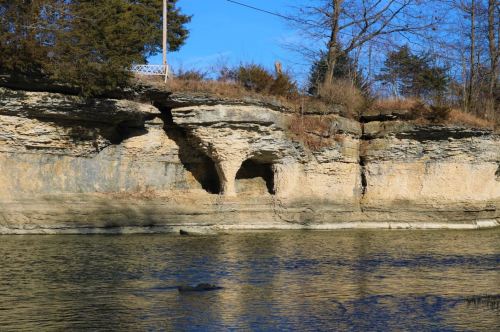 A rocky cliff with caves along a calm river, surrounded by trees under a clear blue sky.