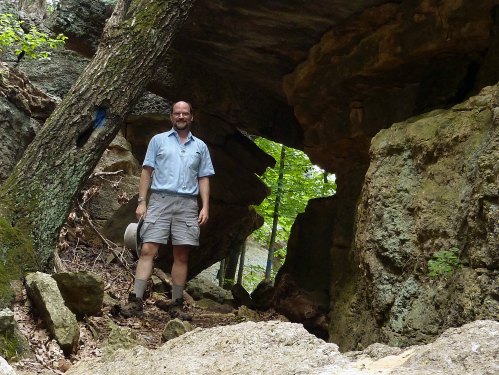A man stands in a rocky area with trees, wearing a light shirt and shorts, surrounded by natural rock formations.