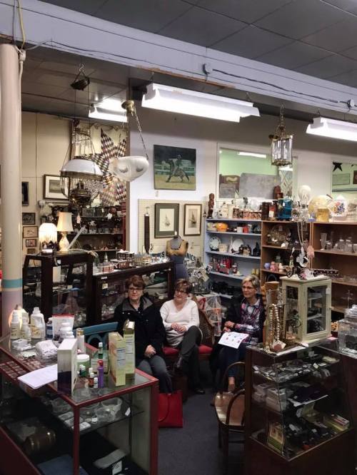 Three women sit in a cozy antique shop surrounded by various vintage items and decor.