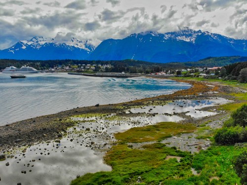 Scenic view of a coastal landscape with mountains, a calm bay, and reflections in the water under a cloudy sky.
