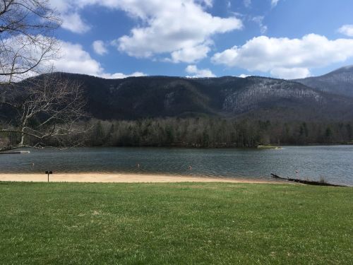 A serene lake surrounded by mountains, with a sandy shore and a clear blue sky dotted with clouds.