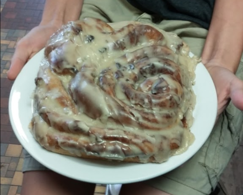 A person holds a large, gooey cinnamon roll on a white plate, topped with creamy icing.