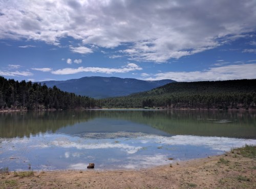 A serene lake surrounded by trees and mountains under a partly cloudy sky, reflecting the landscape in the water.