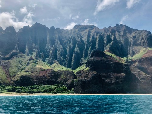 Lush green cliffs rise dramatically above a calm blue ocean, with a sandy beach at the base under a bright sky.