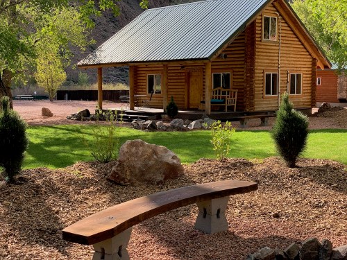 A log cabin with a metal roof sits in a green yard, surrounded by trees and a stone bench in the foreground.