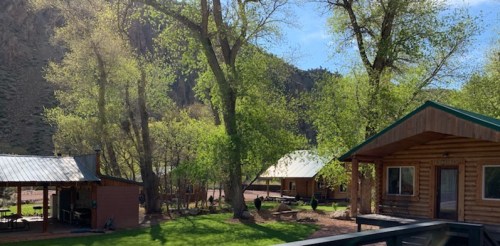 A scenic view of log cabins surrounded by lush green trees and mountains under a clear blue sky.
