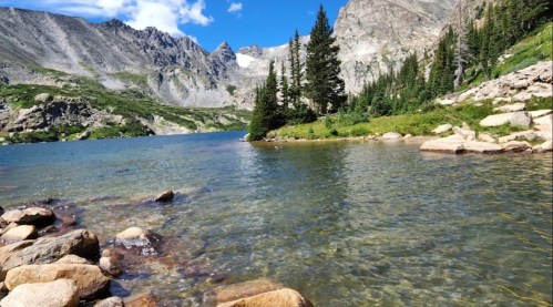 A serene lake surrounded by rocky shores and tall trees, with mountains in the background under a clear blue sky.