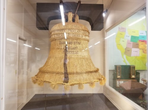 A large straw bell displayed in a glass case, featuring inscriptions and surrounded by a museum setting.