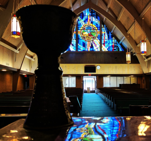 A chalice in the foreground with a colorful stained glass window and empty church interior in the background.