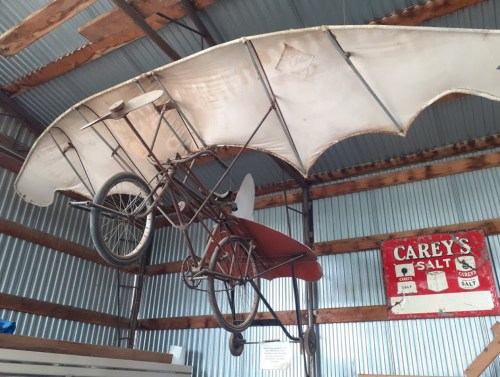 A vintage flying machine made from a bicycle and fabric wings, displayed in a rustic barn with a salt sign in the background.