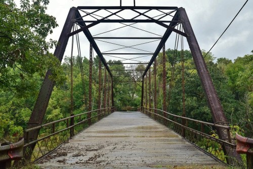 An old metal bridge with a wooden deck, surrounded by lush green trees under a cloudy sky.
