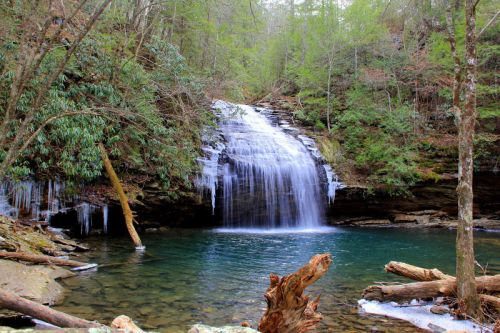A serene waterfall cascades into a clear pool, surrounded by lush greenery and icicles hanging from rocks.