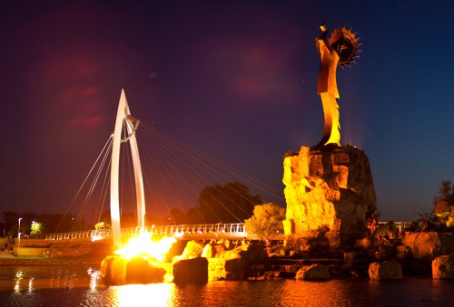 A illuminated statue on a rock beside a bridge at night, reflecting in the water with a dark sky above.