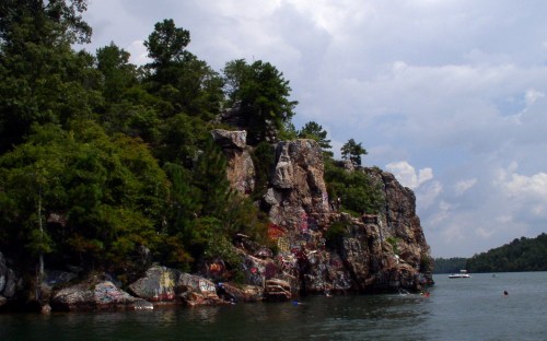 A rocky cliff covered in greenery and graffiti, with people swimming nearby in a calm lake under a cloudy sky.