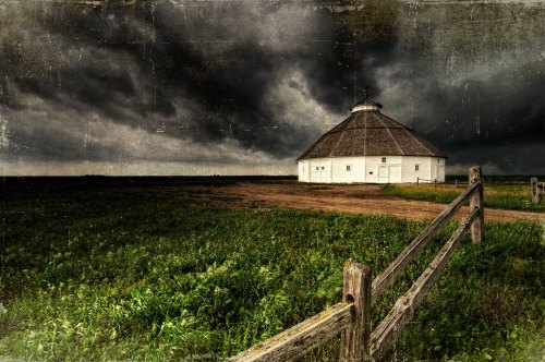 A round white barn stands in a grassy field under a dark, stormy sky, with a wooden fence in the foreground.