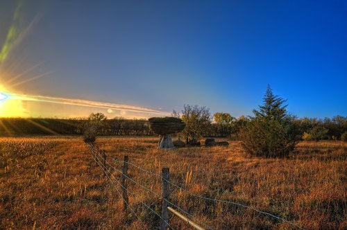A serene landscape at sunset featuring a large rock formation, grassy fields, and a clear blue sky.