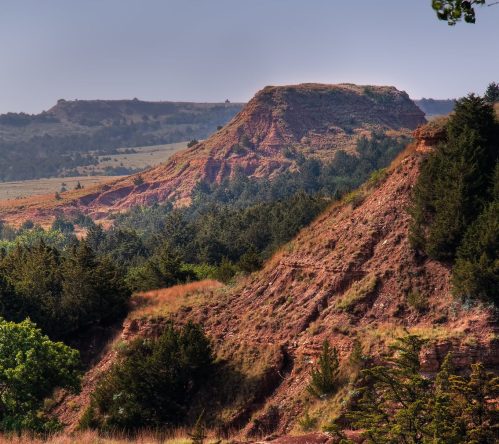 A scenic view of rugged red hills and green trees under a clear sky, showcasing a natural landscape.