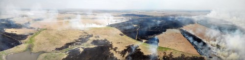 Aerial view of a landscape affected by fire, showing blackened earth and smoke rising over fields and water.