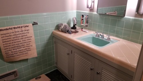 A vintage bathroom with mint green tiles, a sink, and a towel displaying "Wonderful World of Color" information.