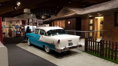 A vintage two-tone car displayed in a museum, with exhibits and a model house in the background.