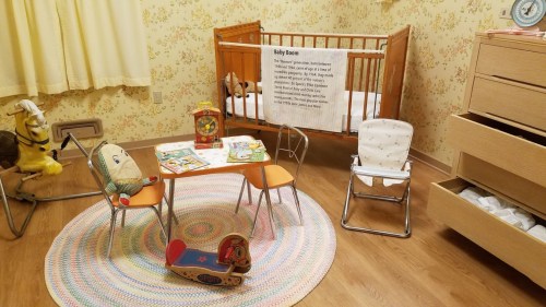 A cozy nursery with a crib, toy chairs, a play table, and a colorful rug, featuring vintage toys and floral wallpaper.