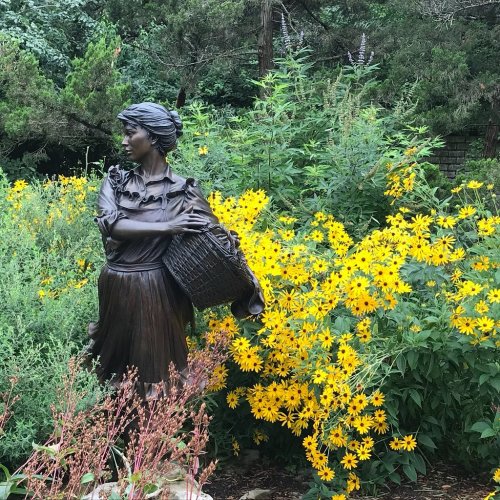 A bronze statue of a woman holding a basket, surrounded by vibrant yellow flowers and lush greenery.