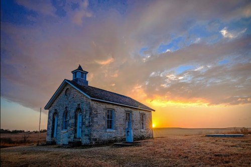 A historic stone building at sunset, with a colorful sky and open fields in the background.