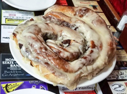 A large, spiral cinnamon roll topped with thick white icing on a plate, resting on a colorful table surface.