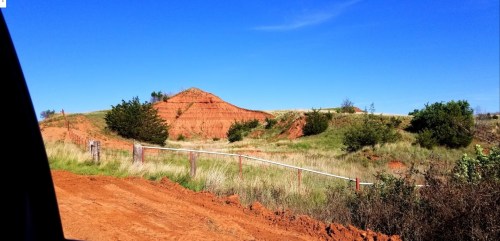 Red dirt landscape with a small hill, green shrubs, and a clear blue sky in the background.
