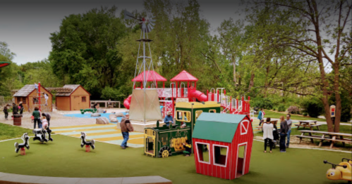 A colorful playground with a train structure, slides, and playhouses, surrounded by trees and families enjoying the space.