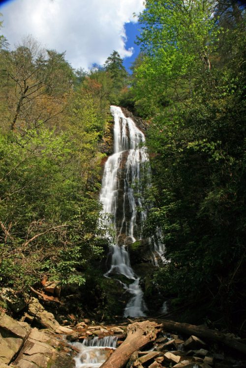 A tall waterfall cascades down rocky terrain, surrounded by lush green trees and a bright blue sky.