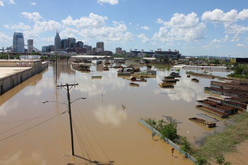 A flooded urban area with submerged buildings and debris, against a backdrop of a city skyline and blue sky.