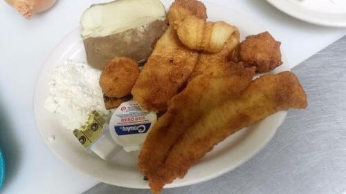 A plate of fried fish fillets, hushpuppies, a baked potato, and a side of cottage cheese with condiments.