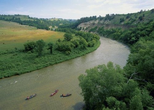 Aerial view of a winding river surrounded by lush greenery and hills, with kayakers paddling along the water.